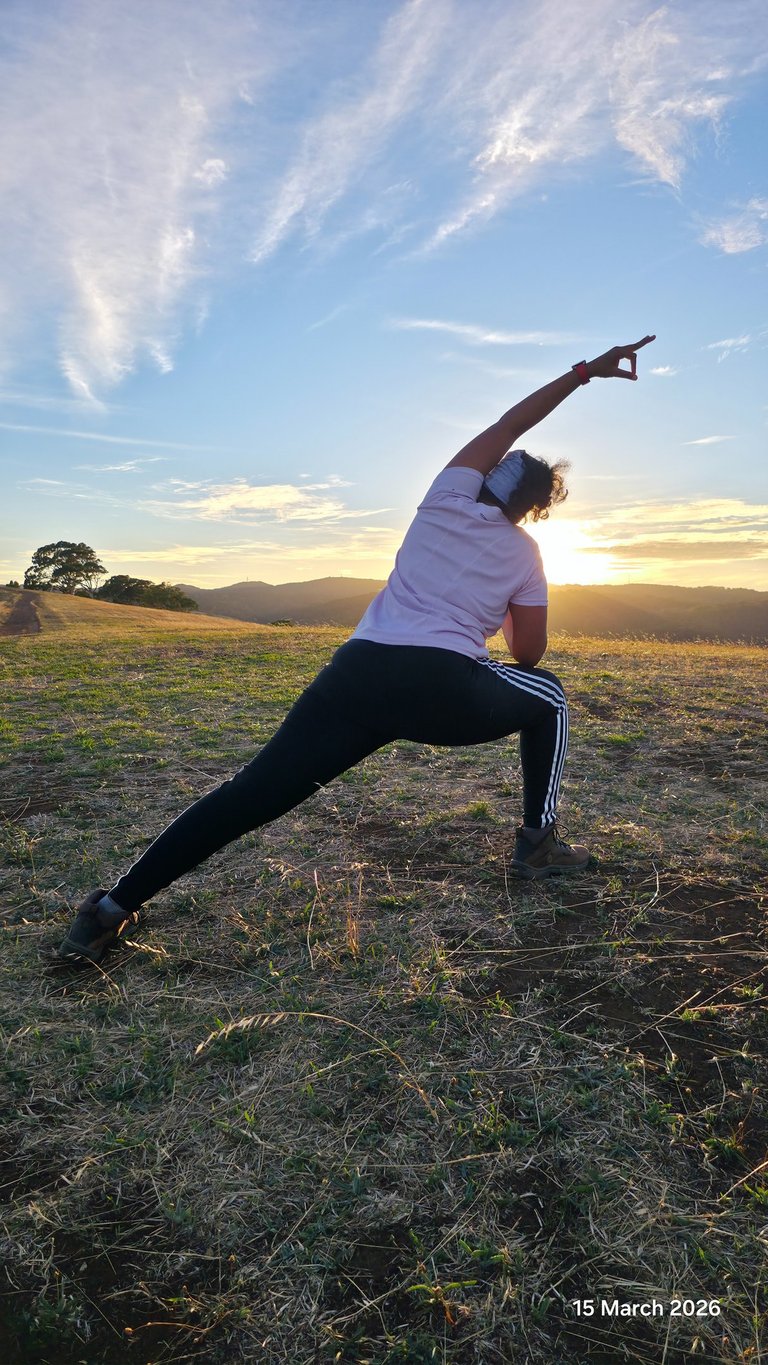 A woman practices outdoor yoga in a side lunge pose at sunset on a grassy hilltop.