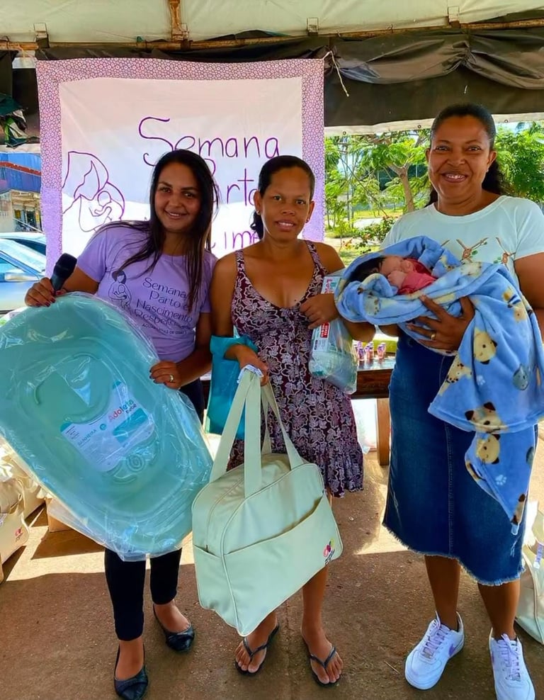 Three women holding baby care gifts and a newborn at a Semana do Parto community event.