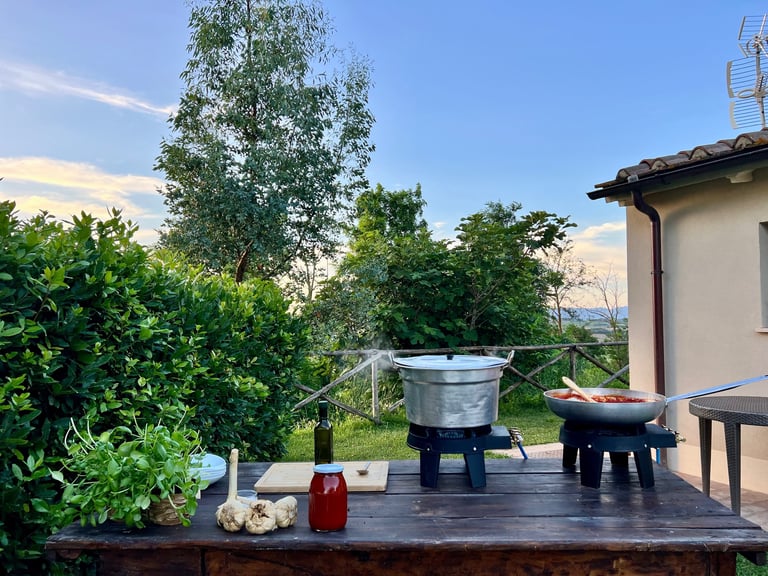 Outdoor cooking setup with large pots on gas burners, fresh herbs, and garlic on a wooden table.