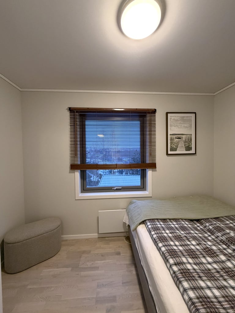 Cozy minimalist bedroom with a plaid duvet, wooden window blinds, and a gray ottoman on light wood floors.