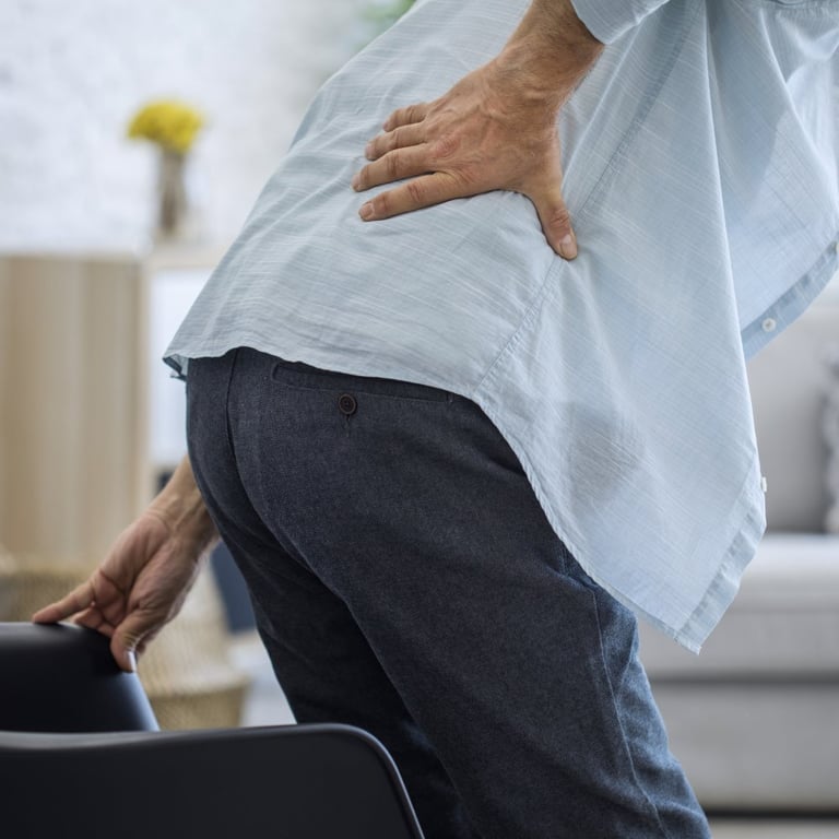 A man holding his lower back in pain while standing up from a chair, highlighting symptoms of sciatica or back strain.