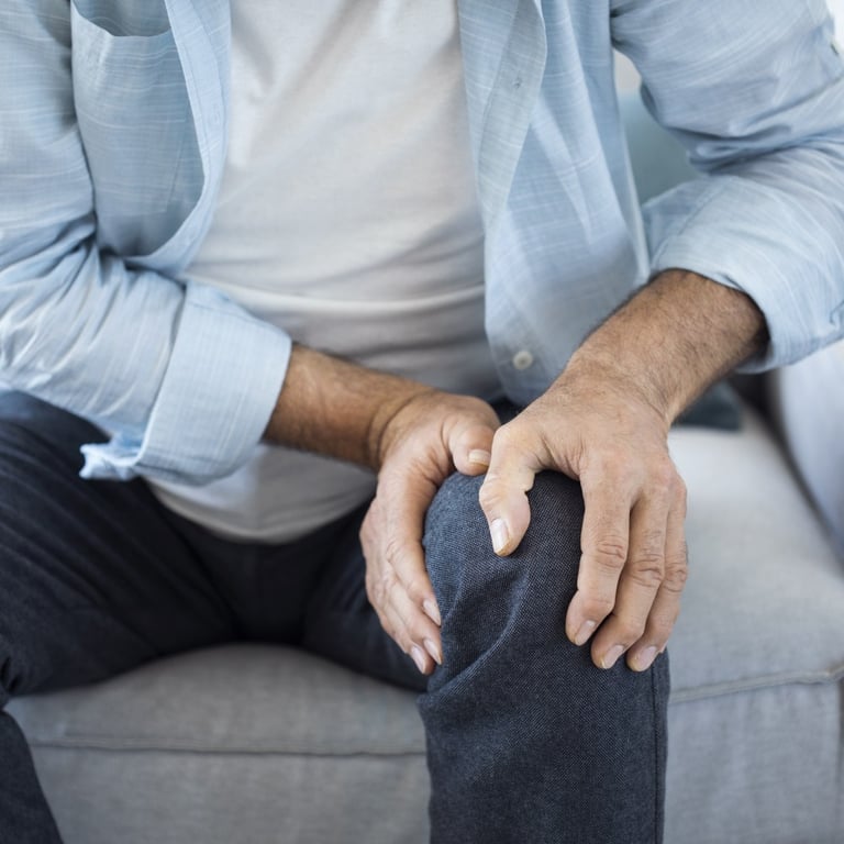 Close-up of a man sitting on a sofa and holding his painful knee due to joint inflammation or arthritis.