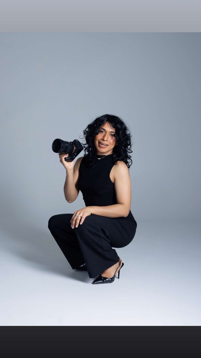 A smiling female photographer with curly hair holding a professional DSLR camera in a studio.