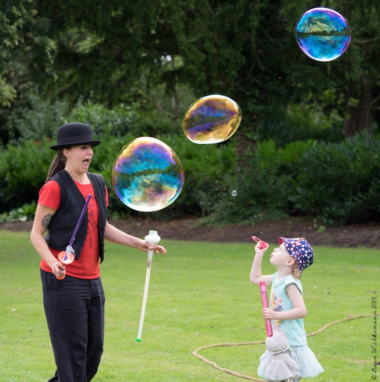 A woman and young child blowing giant iridescent soap bubbles in a green park.