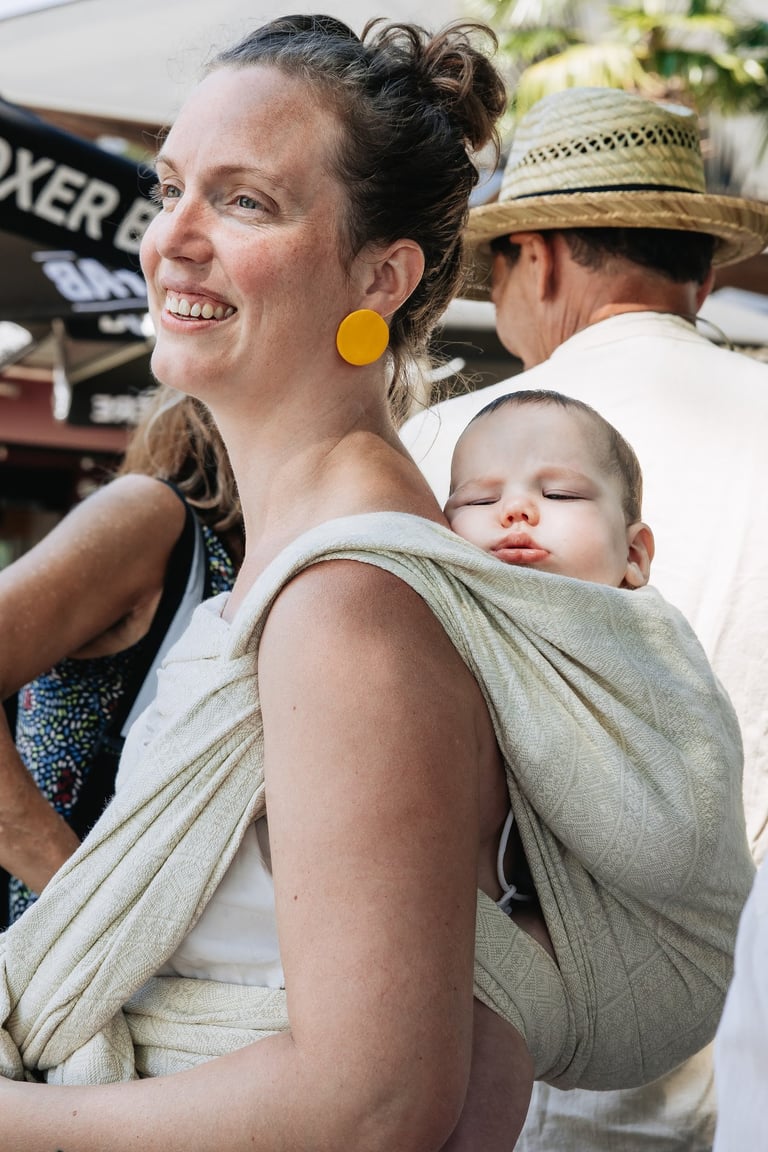 Smiling woman wearing a sleeping infant in a woven linen baby wrap carrier at an outdoor market.
