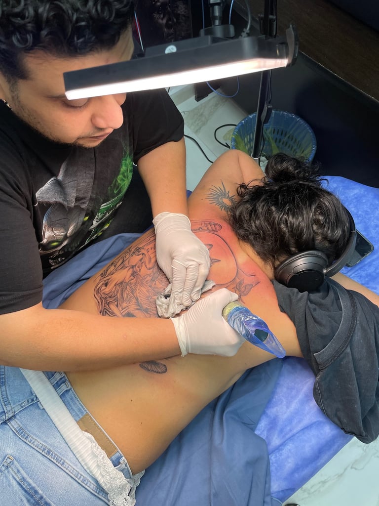 Tattoo artist working on a large back tattoo using a rotary machine under LED lighting in a Hurghada