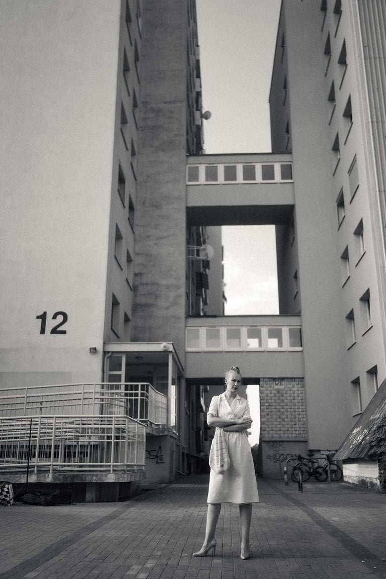 A black and white photo of a woman in a dress standing before a brutalist apartment building.