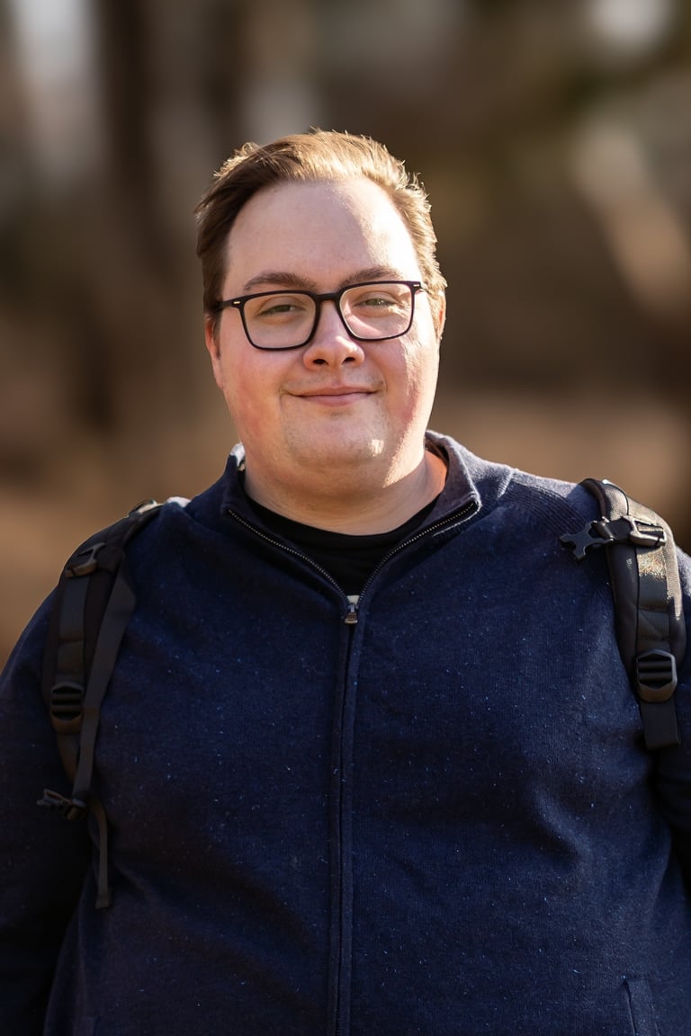 A smiling man with glasses wearing a blue zip-up sweater and a backpack outdoors.