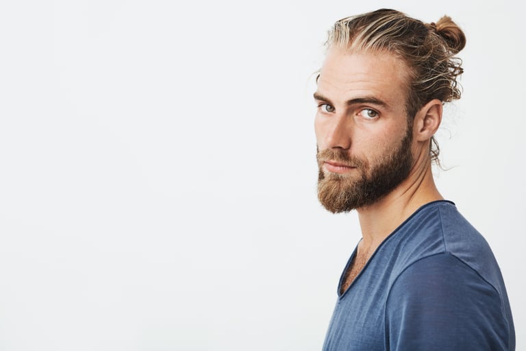 Handsome man with a blonde man bun and groomed beard posing against a white background.