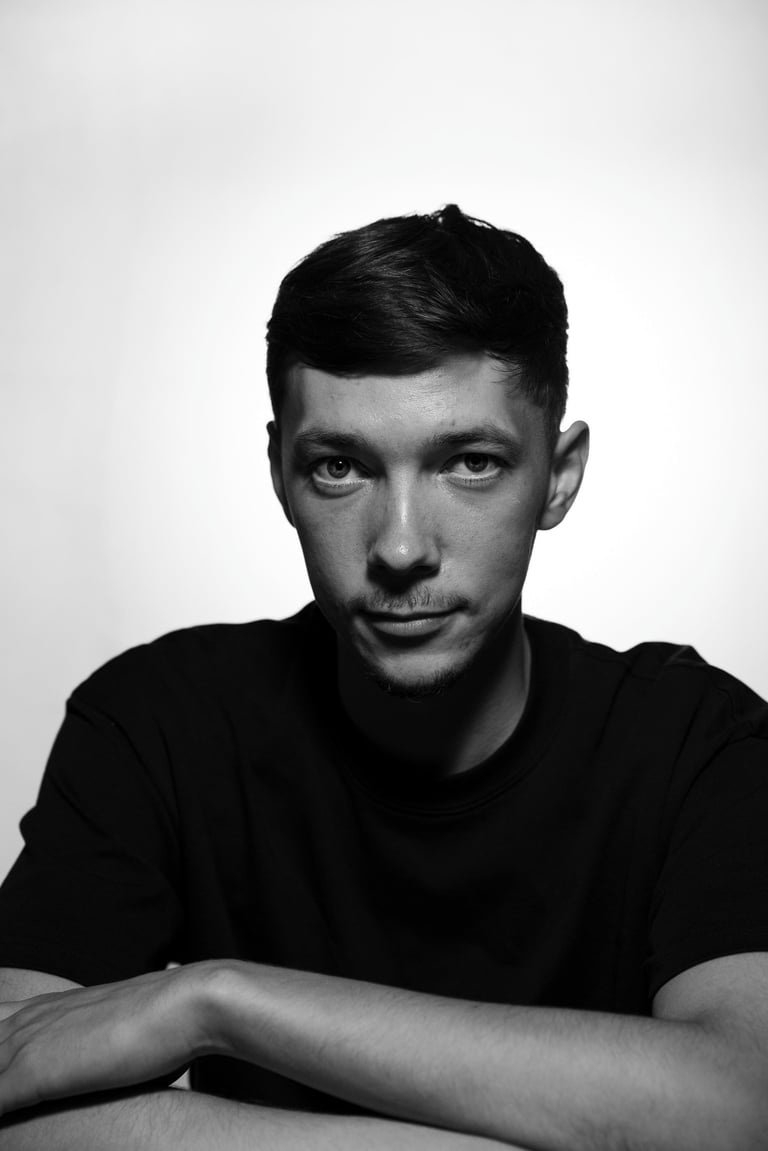 Black and white portrait of a young man with dark hair in a casual black t-shirt against a white background.