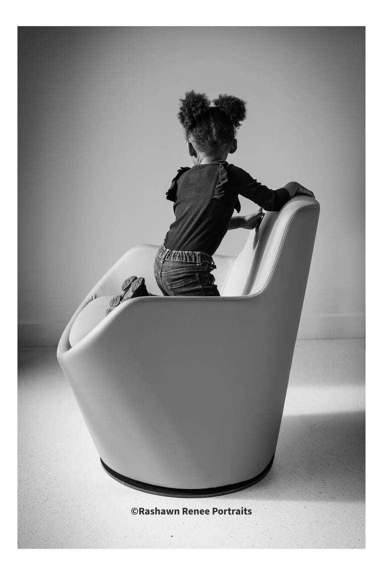 Black and white portrait of a young girl with afro puffs climbing on a modern studio chair.