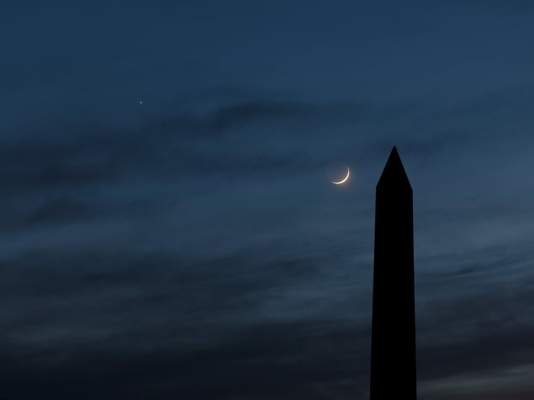 Silhouette of a stone obelisk against a dark blue twilight sky with a delicate crescent moon and a bright planet.