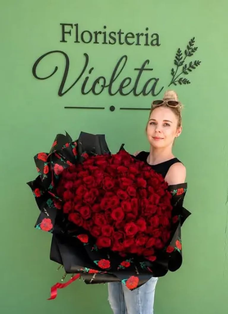 A woman holds a massive bouquet of red roses in front of a green wall with the sign Floristeria Violeta. Delivery Time