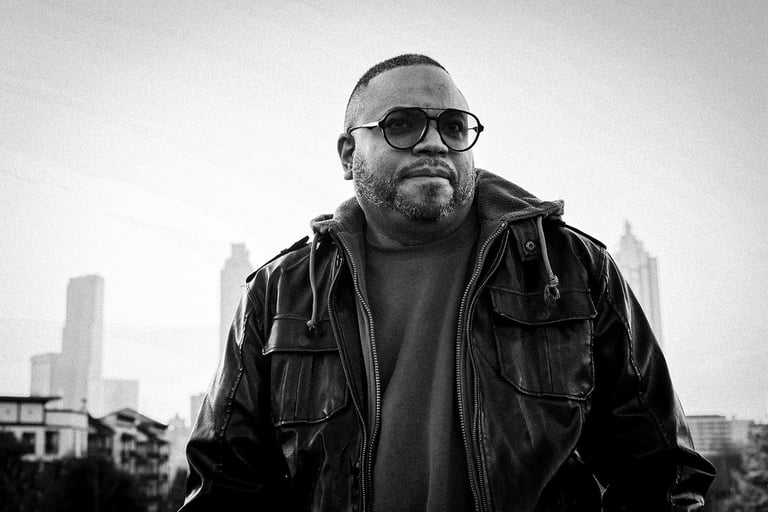 Portrait of Rico Sanchez in a leather jacket and sunglasses with a city skyline background.