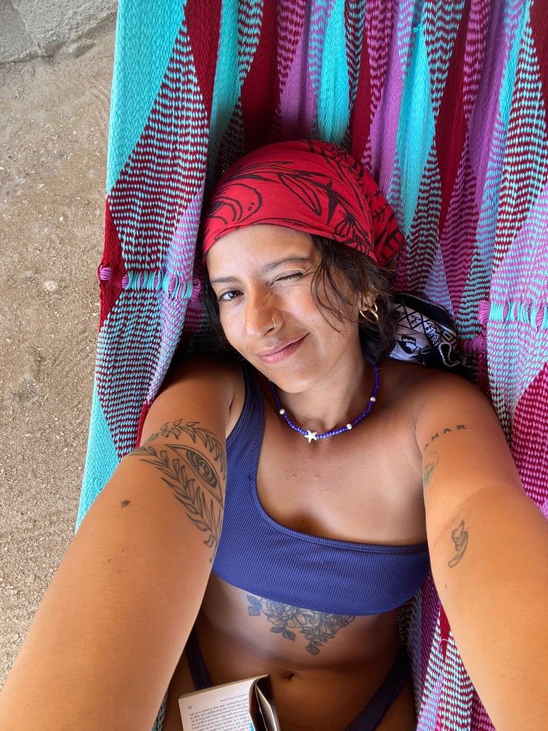 Mujer tatuada con un pañuelo rojo guiñando el ojo mientras se relaja en una colorida hamaca afuera.