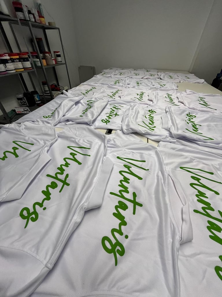 Custom screen printed white volunteer t-shirts with green cursive text laid out in a workshop.