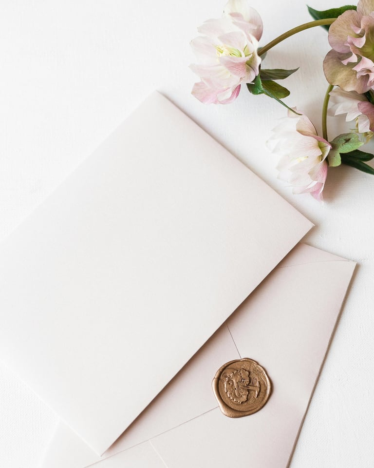 Elegant blank invitation card and envelope with gold wax seal next to pink hellebore flowers.