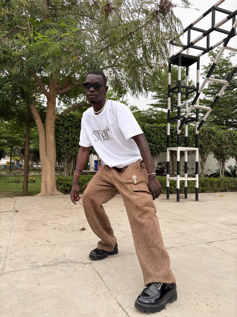 A man in a mountfit white graphic t-shirt and brown cargo pants posing in a park with sunglasses.