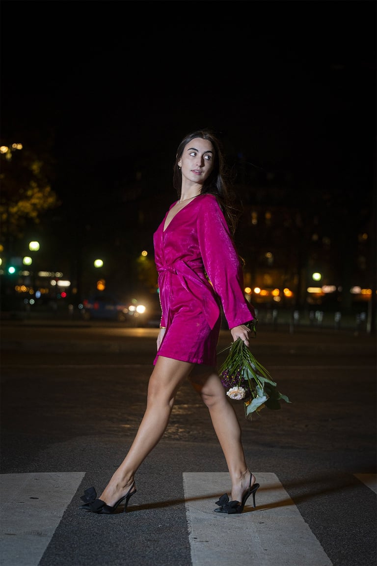 Femme en robe fuchsia traversant un passage clouté de nuit à Paris, bouquet à la main. Photo par le photographe Cyril Steau. 