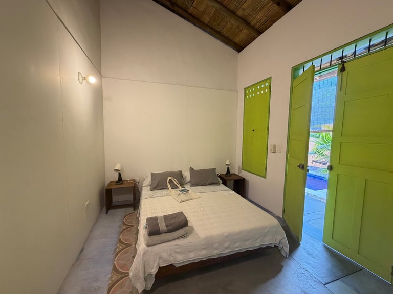 Rustic tropical bedroom with white bedding, wooden ceiling, and bright green shutters and door.