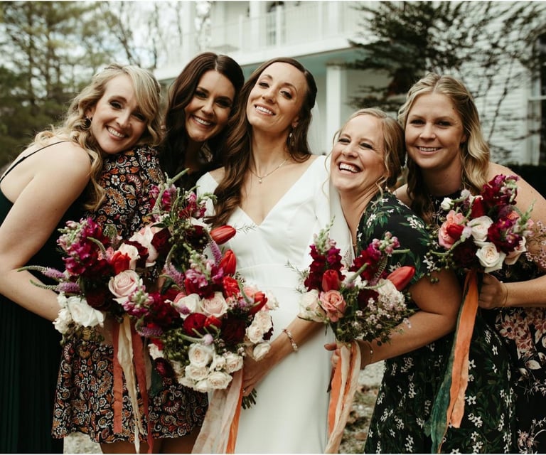 Smiling bride and bridesmaids holding red and pink floral bouquets at a wedding celebration.