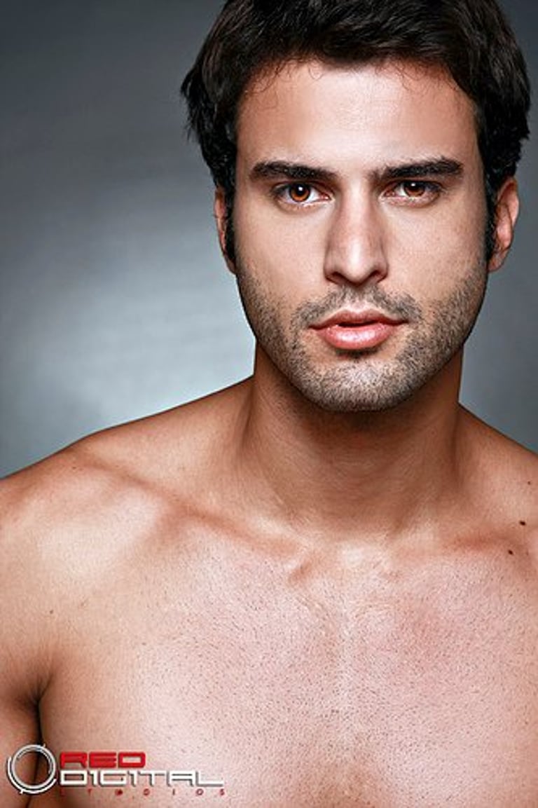 Close-up headshot of a male model with brown eyes and short stubble beard against a gray background.