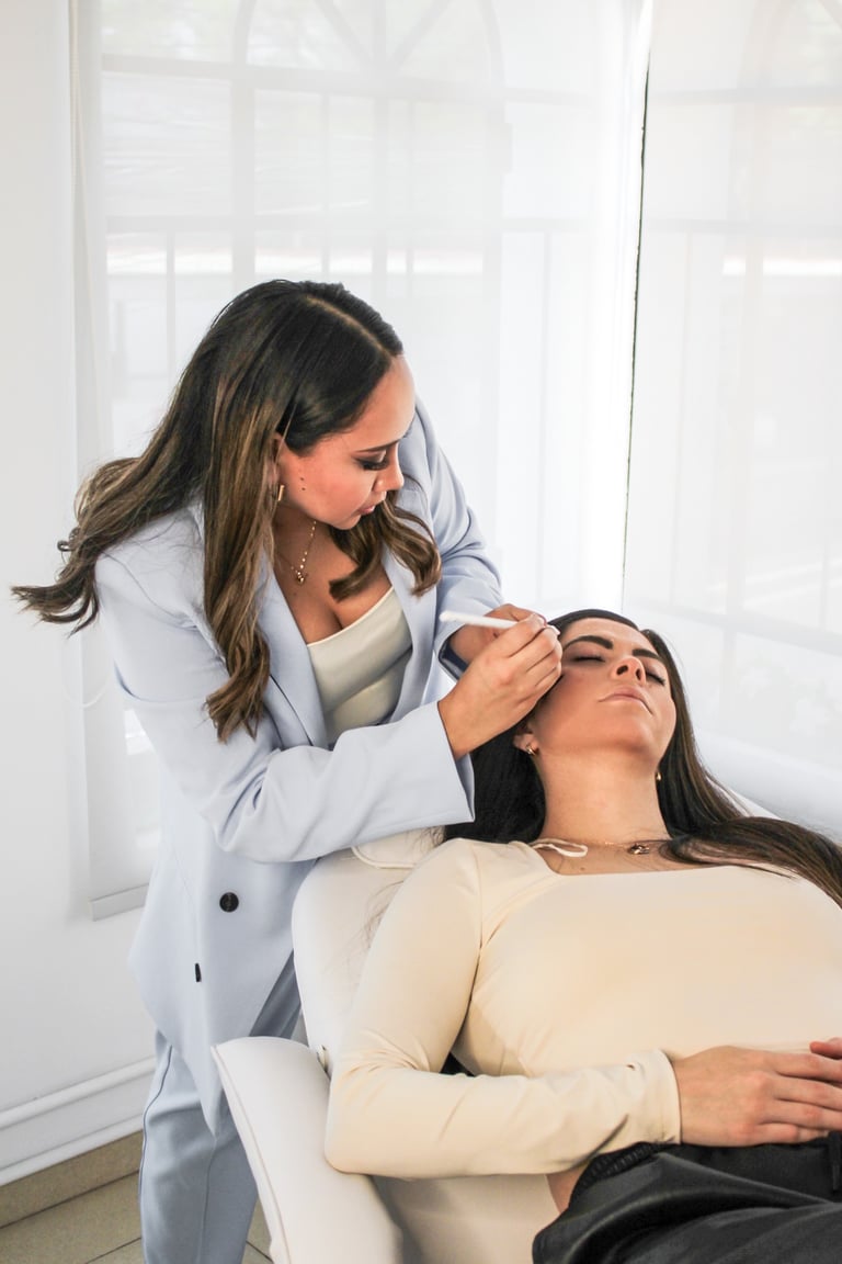 A professional brow artist performs microblading mapping on a client's eyebrows in a bright studio.