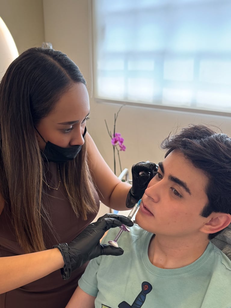 A medical professional in a face mask provides facial filler injections to a male patient's jawline.