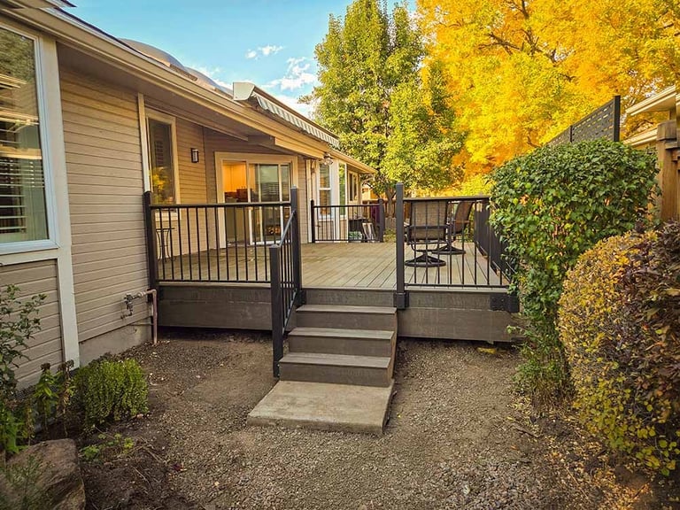 Residential house composite deck with black metal railings, wooden stairs, and a dirt yard. Autumn trees with yellow leaves.