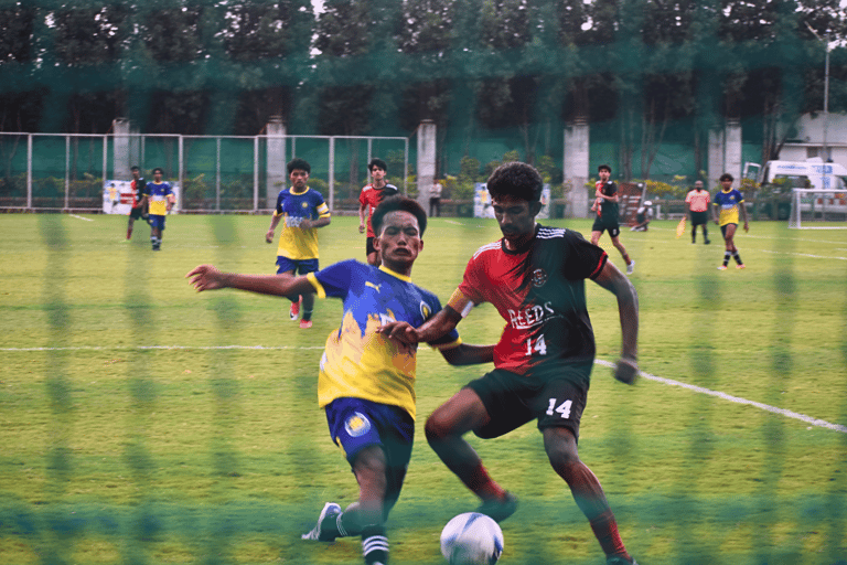 Two soccer players in red and blue jerseys compete for the ball during a competitive match on a grass field.