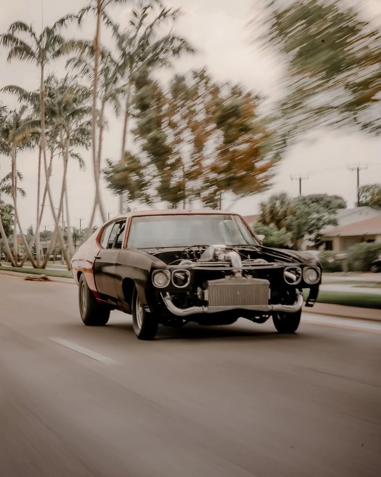 Custom black and red muscle car with exposed turbo engine driving fast past tropical palm trees.