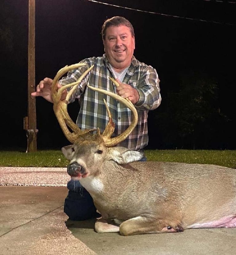 Owner guide kneels beside and holds up the large antlers of a harvested whitetail deer at night on a paved surface.