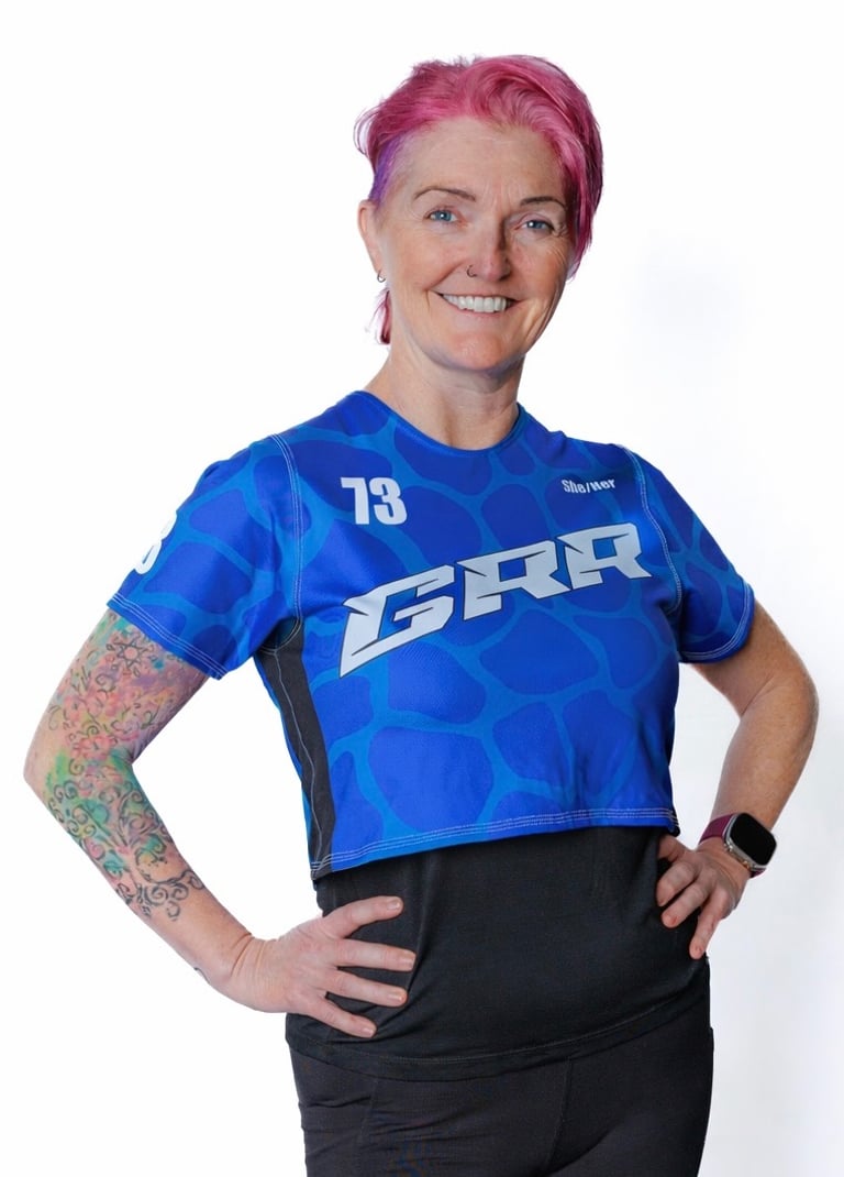 Smiling roller derby athlete with pink hair wearing a blue GRR team jersey and colorful arm tattoo.