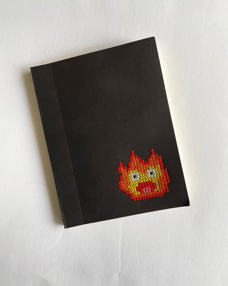 Cuaderno negro con una llama de Calcifer bordada a mano en punto de cruz en la portada.