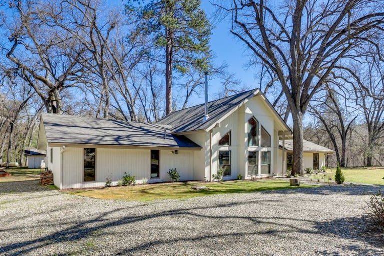 Modern white ranch house with large windows and vaulted ceilings surrounded by tall oak trees.