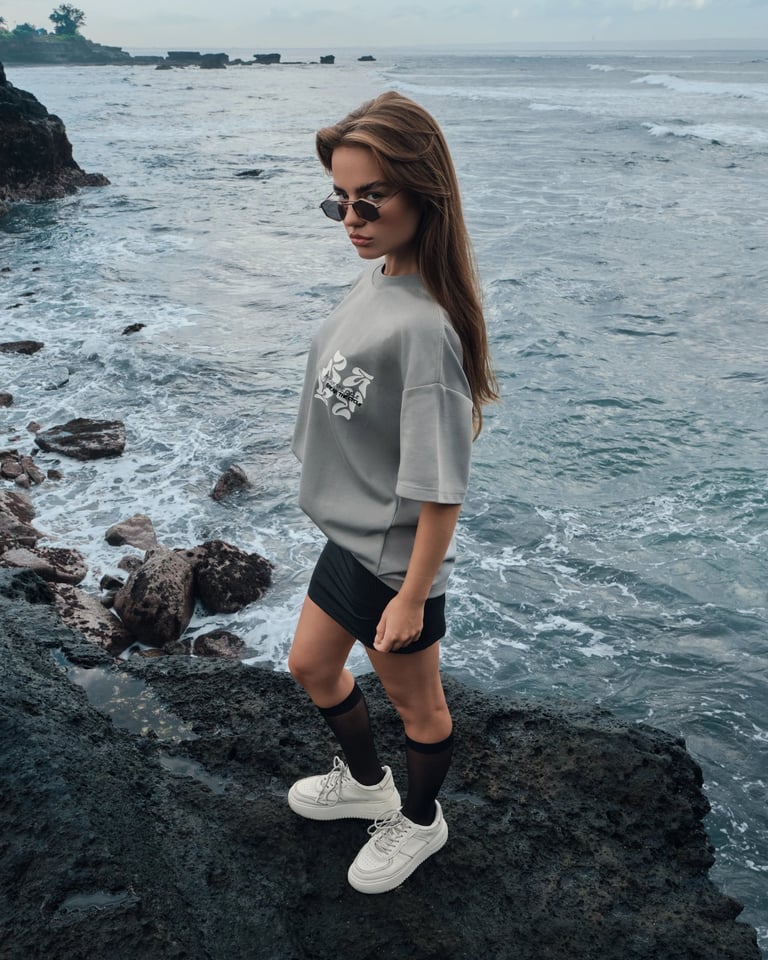 Model in gray oversized t-shirt and miniskirt standing on volcanic cliff coast for Break the Cycle summer campaign 2025.