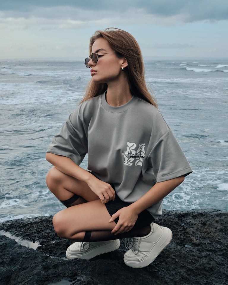 Model in gray oversized t-shirt and miniskirt sitting on volcanic cliff coast for Break the Cycle summer campaign 2025.