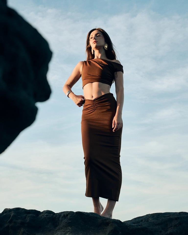 Model in brown dress on volcanic coastline rocks, Bali fashion editorial.