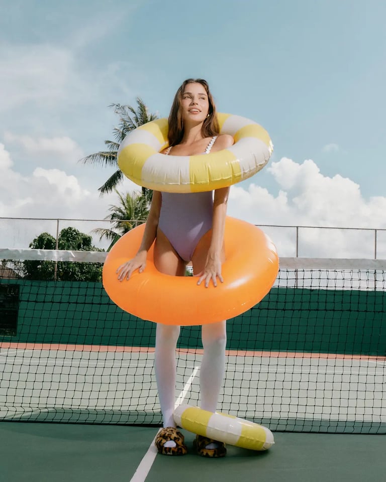 Model in purple swimsuit inside a yellow-orange float, shot on a tennis court with bright ambient light.