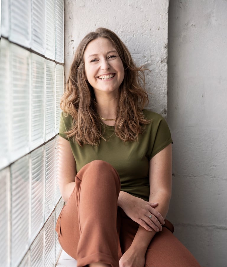 Smiling woman in olive green t-shirt sitting by a glass block window for a professional headshot.