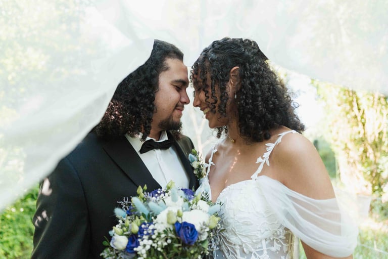Un couple s'apprête à s'embrasser sous un voile flottant, la mariée tenant un bouquet bleu et blanc.