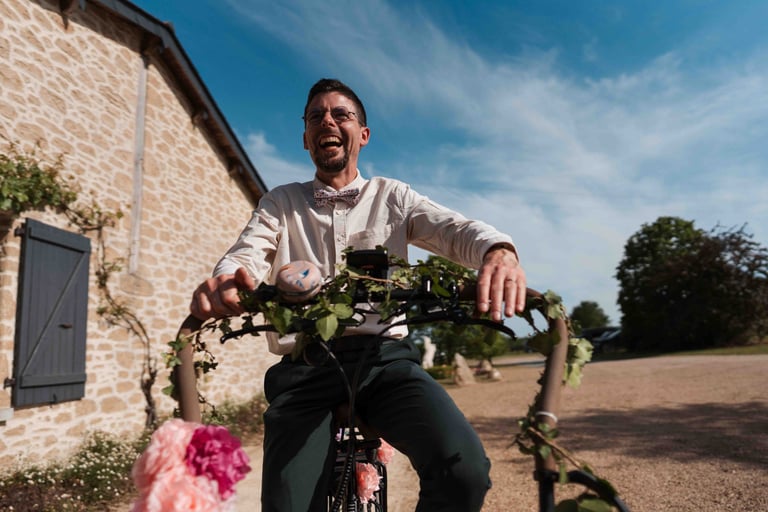 Un marié souriant, à vélo décoré de fleurs, devant un bâtiment rustique en pierre, par une journée de mariage ensoleillée.