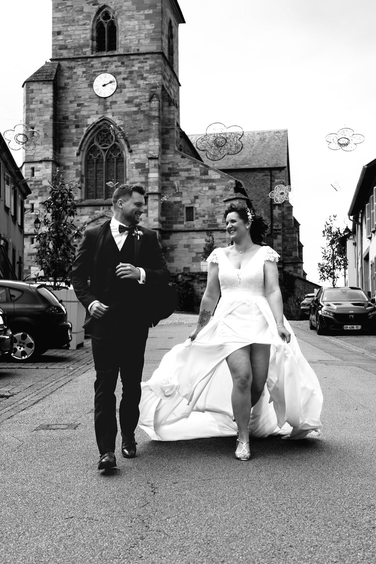 Photo en noir et blanc d'un couple de mariés souriants marchant devant une église en pierre