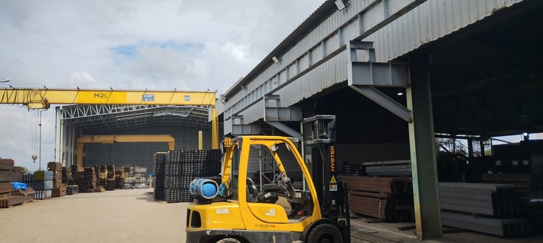 Empilhadeira amarela em frente a galpão industrial com estrutura metálica, guindaste e materiais.