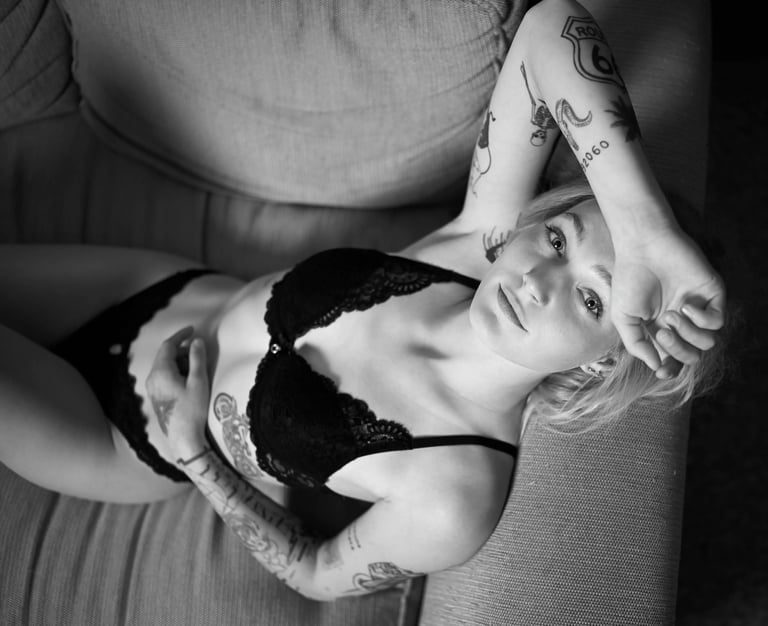 Schwarz-Weiß-Boudoirfoto einer tätowierten Frau in schwarzer Spitzenlingerie, die entspannt auf einem Sofa liegt; ästhetische