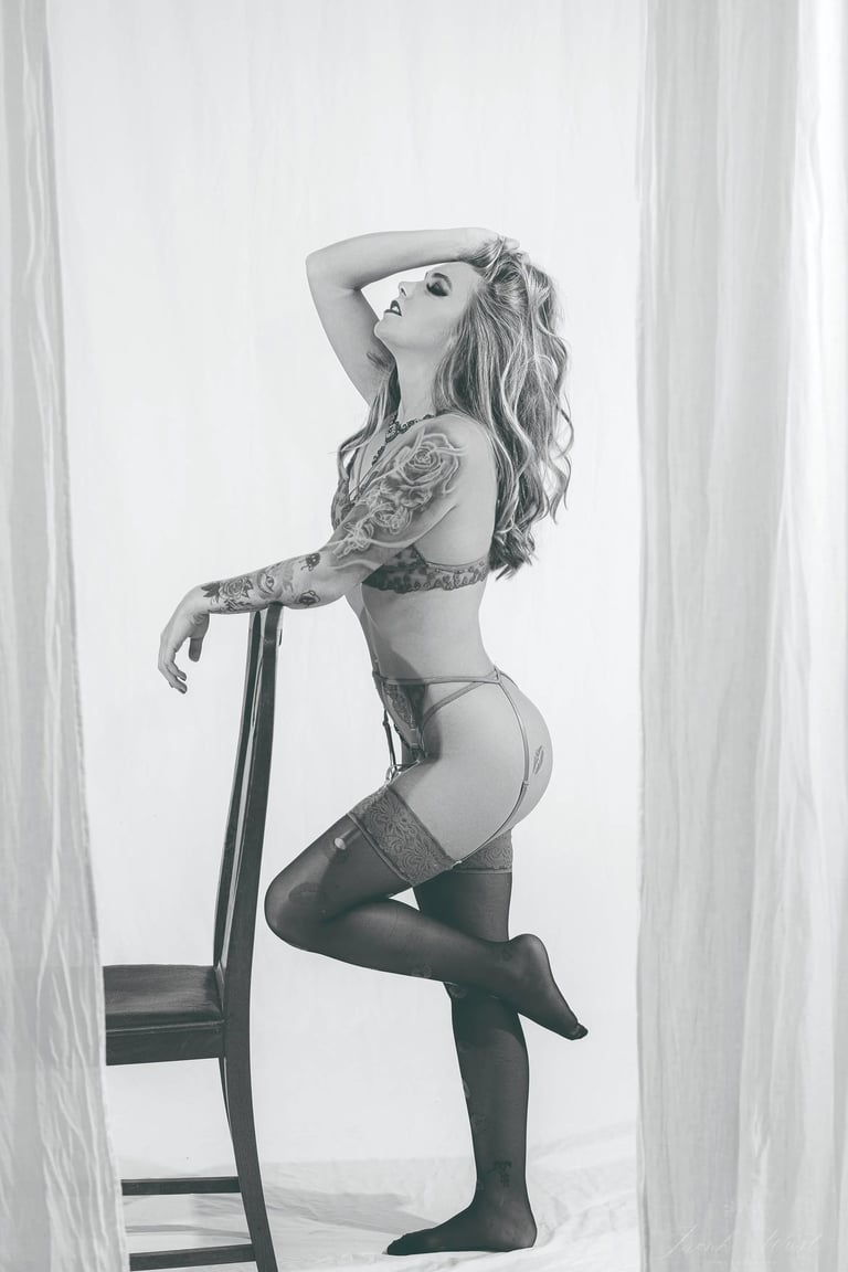 Schwarz-Weiß-Boudoirfoto einer tätowierten Frau in Lingerie, die elegant an einem Stuhl posiert; ästhetisches Fineart-Portrai