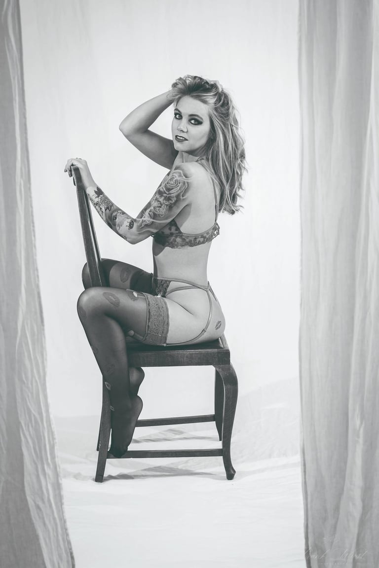 Schwarz-Weiß-Boudoirfoto einer tätowierten Frau, die in Lingerie seitlich auf einem Stuhl sitzt und zur Kamera blickt; ästhet