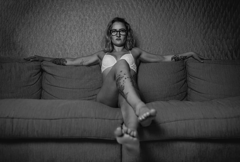 Eine Frau sitzt selbstbewusst auf einem Sofa, trägt lediglich einen hellen BH und eine Brille. Ihre Arme sind seitlich ausges
