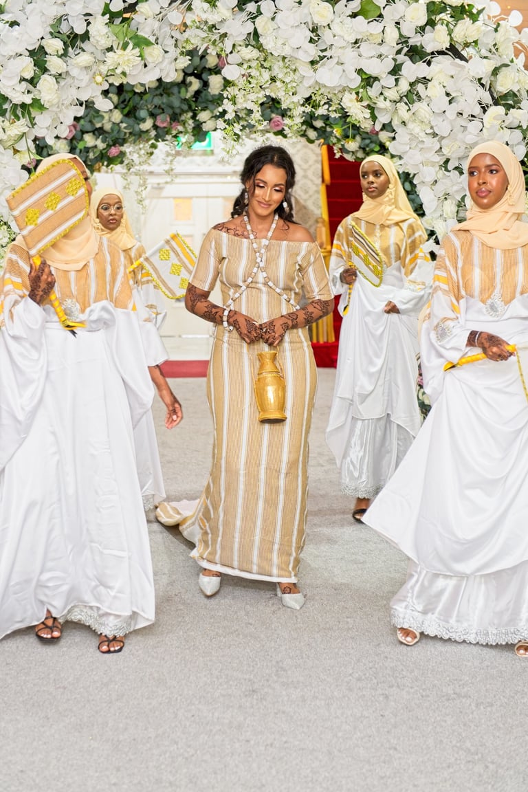 Happy Somalian bride and bridesmaids entering wedding venue all in traditional white & gold dresses, floral arch framing them