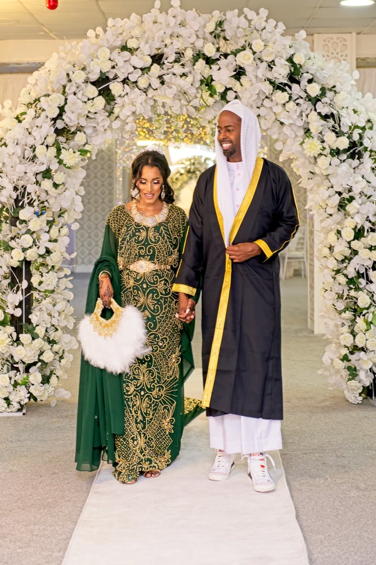 Somalian bride & groom dressed in traditional wedding outfits holding hands and walking, both happy, floral arch framing them